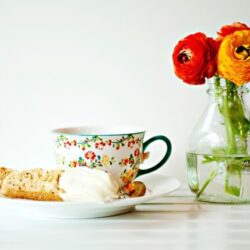 Fresh baked cookie with creamy ice cream, colorful floral mug, and vibrant ranunculus flowers in a glass vase on a white surface.