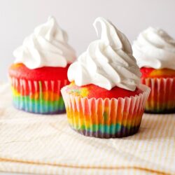 Vibrant rainbow cupcakes with whipped frosting on a textured cloth.