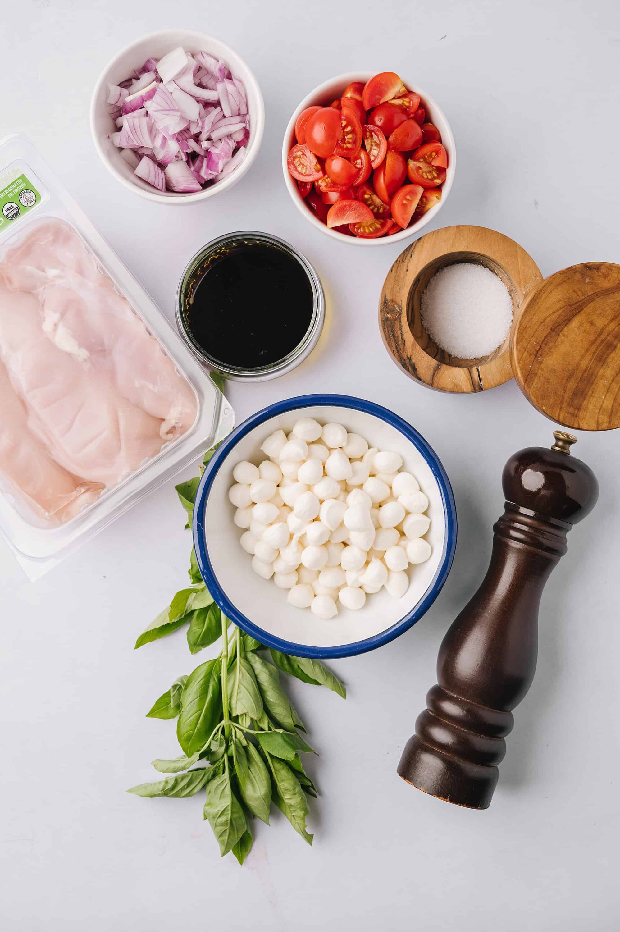 Fresh cherry tomatoes, sliced red onions, mozzarella cheese, basil, balsamic vinegar, salt, and black pepper on a white background.