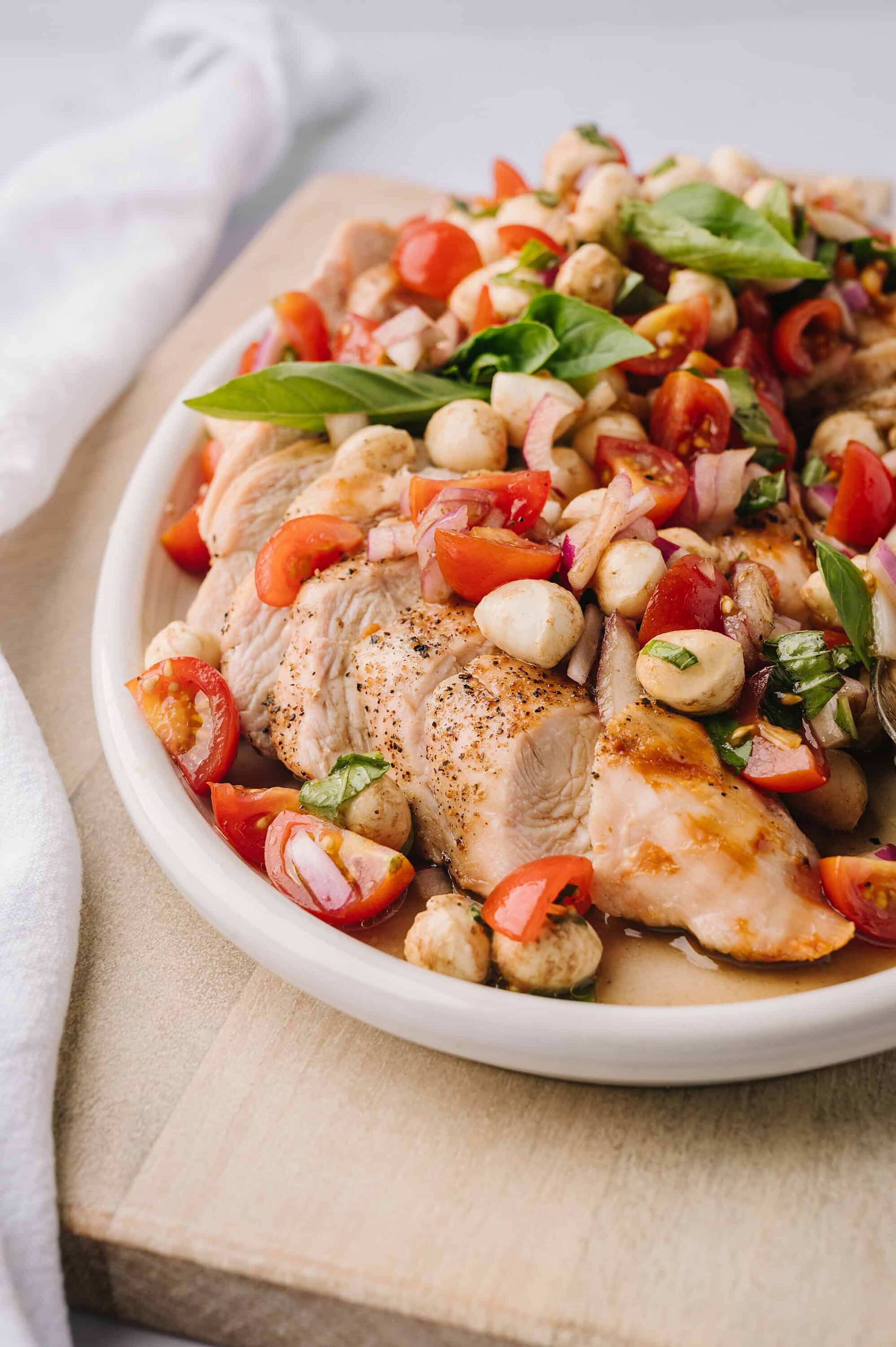 Roasted chicken breast with cherry tomatoes, mozzarella, basil, and onion salad on a white plate.