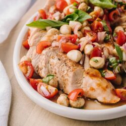 Roasted chicken breast with cherry tomatoes, mozzarella, basil, and onion salad on a white plate.