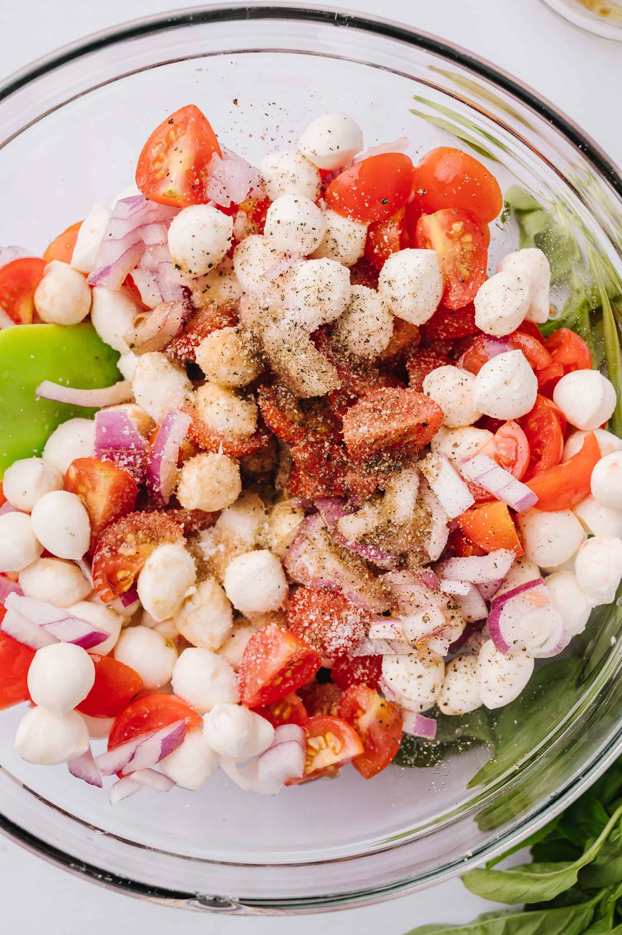 Fresh tomato, mozzarella, and basil salad with red onions and seasoning on a glass bowl, ready for serving.