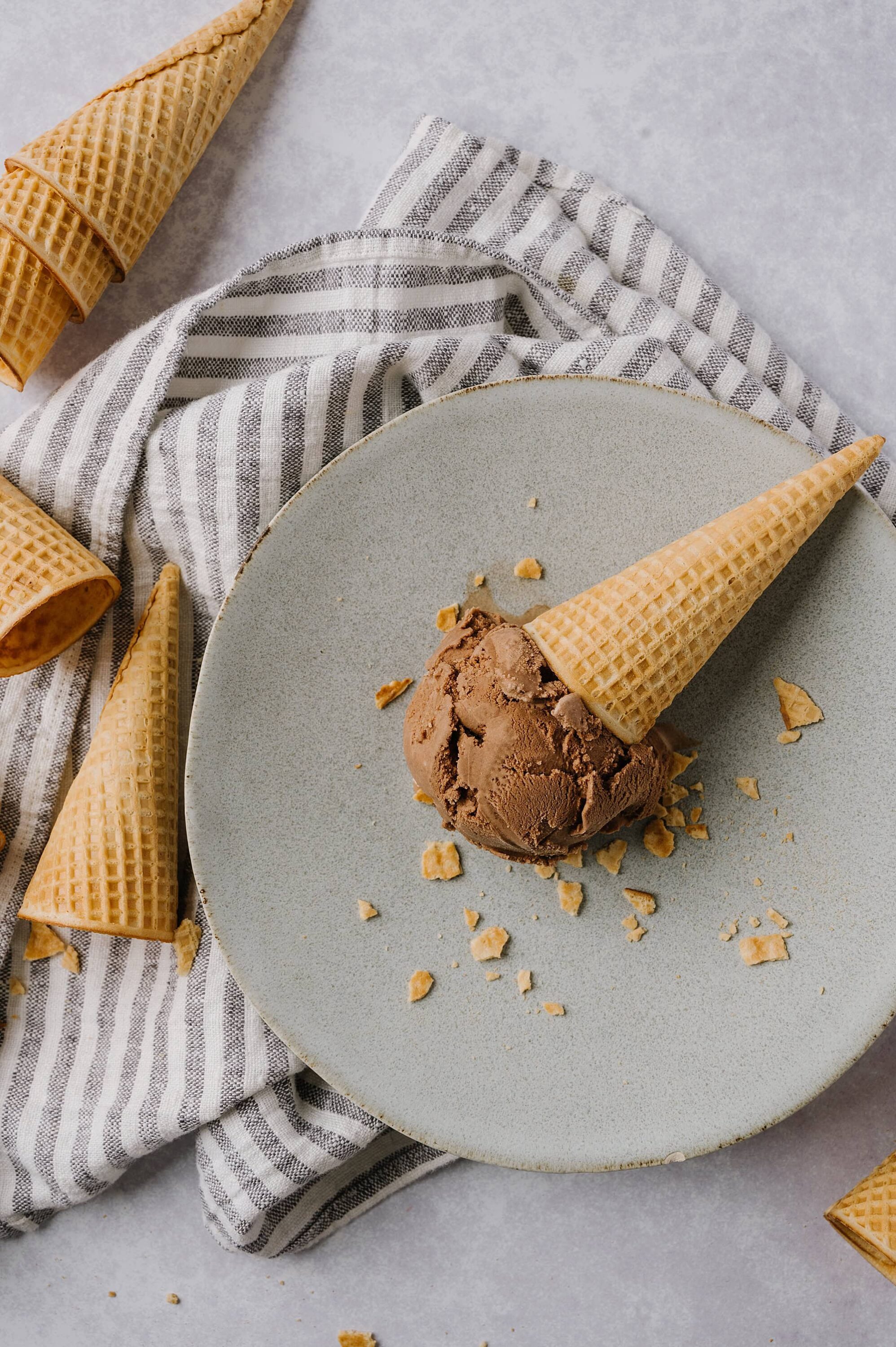 Creamy chocolate ice cream in a waffle cone on a gray plate with scattered cookie crumbs.
