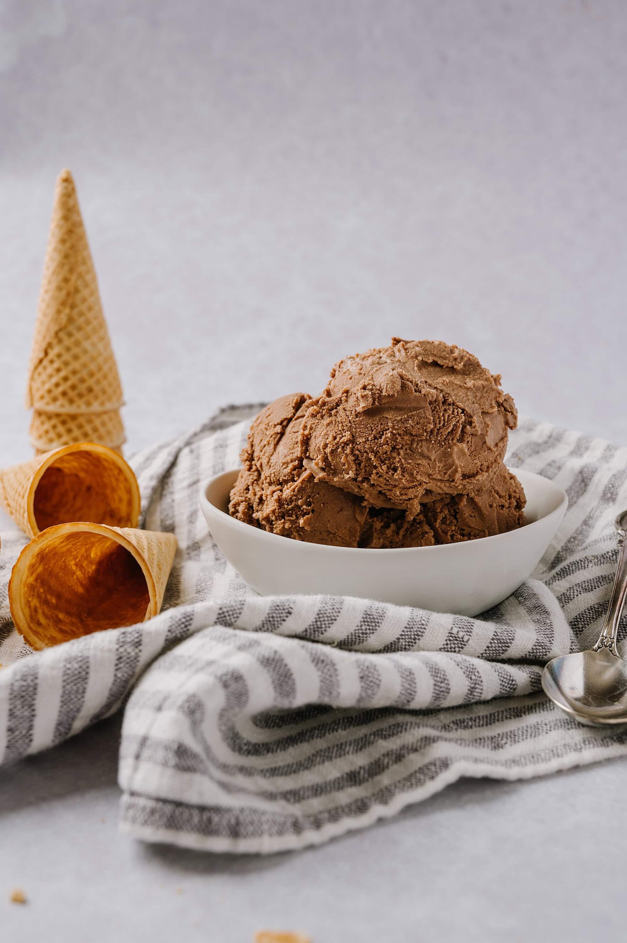 Creamy chocolate ice cream in a white bowl with waffle cones on a striped cloth.
