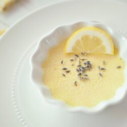 Lemon lavender dessert in a white bowl on a white plate, garnished with lemon wedge and lavender.