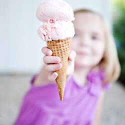 Creamy pink ice cream cone being held by a young girl in a purple shirt.