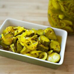 Pickled cucumbers in a white dish with a jar of pickles in the background.