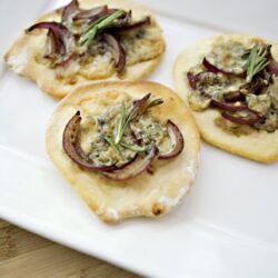 Mini flatbread pizzas topped with red onions, cheese, and herbs on a white serving plate.