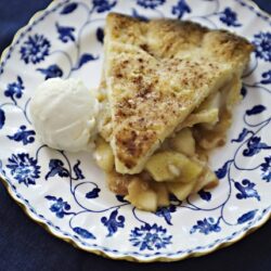 Golden apple cinnamon pie slice with whipped cream on elegant blue and white floral plate.