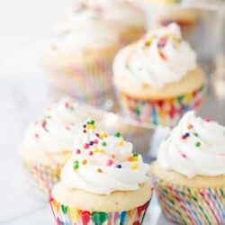 Colorful sprinkles on vanilla cupcakes with swirled frosting, in rainbow cupcake liners.