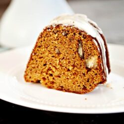 Golden carrot cake slice with cream cheese frosting and crushed pecans on white plate.