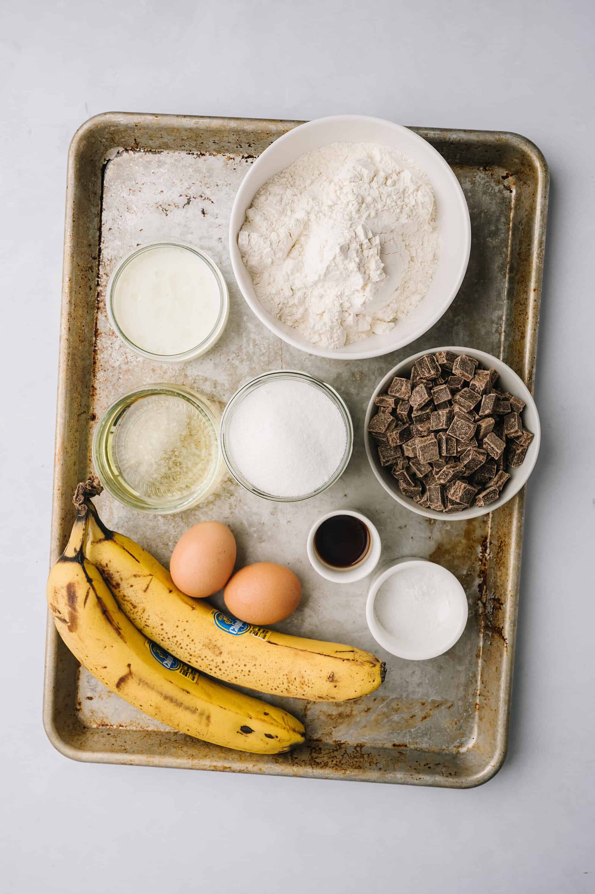 Flour, bananas, eggs, chocolate, and baking ingredients on baking sheet for banana bread.