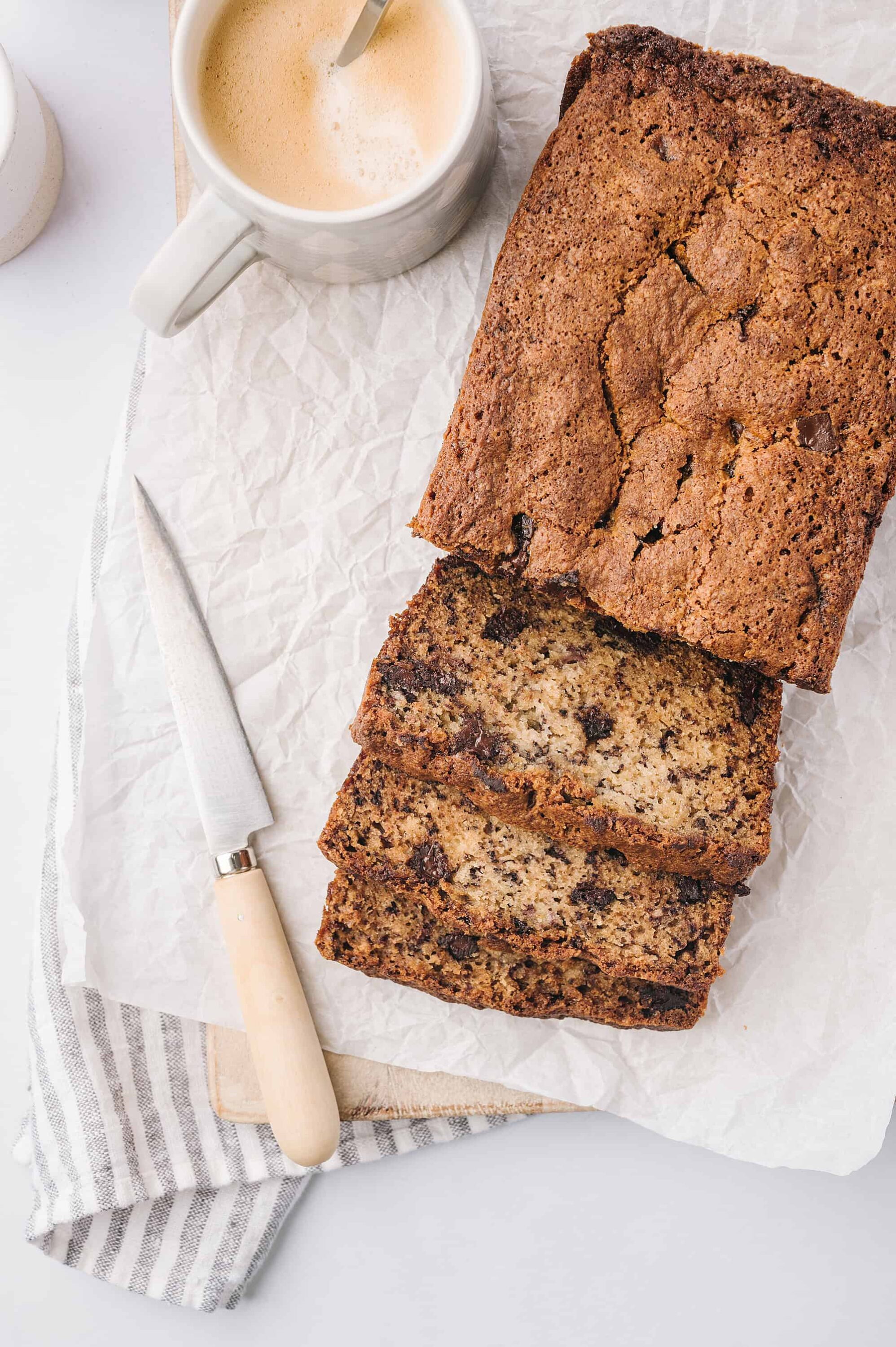 Moist chocolate chip banana bread loaf on parchment paper with a cup of coffee; baked Bree recipe.