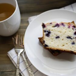 Fresh blueberry loaf cake on a white plate with a cup of tea on a rustic wooden table.