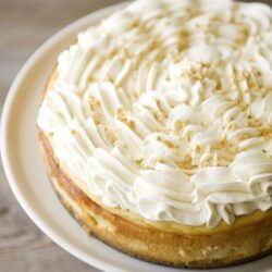 Cream cheese frosting topped cheesecake with white chocolate shavings on a white plate.