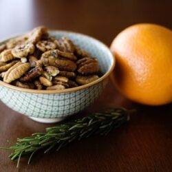 Crunchy toasted pecans in a patterned bowl with fresh orange and sprig of rosemary on a wooden table.