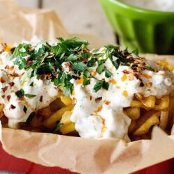 Crispy fries topped with creamy cheese sauce, fresh herbs, and spicy seasonings in a red tray.