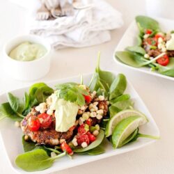 Fresh grilled steak salad with cherry tomatoes, corn, spinach, and lime wedges on a white plate.