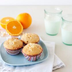 Freshly baked lemon muffins on a blue plate with oranges and glasses of milk.