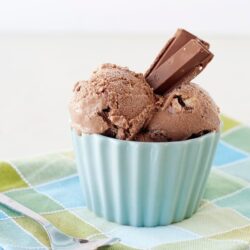 Creamy chocolate ice cream with a chocolate bar garnish in a light blue bowl on a colorful checkered napkin.