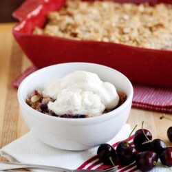Sweet cherry crisp with vanilla ice cream served in a white bowl on a wooden table.