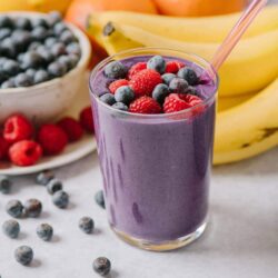 Fresh blueberry smoothie topped with mixed berries and surrounded by bananas, oranges, and blueberries on a white table.