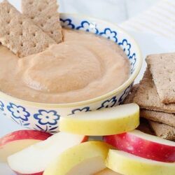 Creamy peanut butter dip with apple slices and whole grain crackers on a white plate, healthy snack idea.