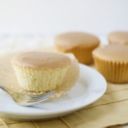 Rich vanilla cupcake with smooth frosting on a white plate, fresh baked cupcakes on a wooden surface.