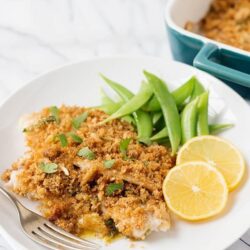 Baked cod with lemon, green beans, and breadcrumb topping on white plate.