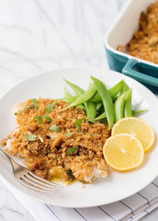 Golden baked fish with lemon, green beans, and breadcrumb topping on a white plate.