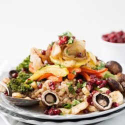 Roasted vegetable grain salad with pomegranate seeds and fresh herbs on a glass plate.