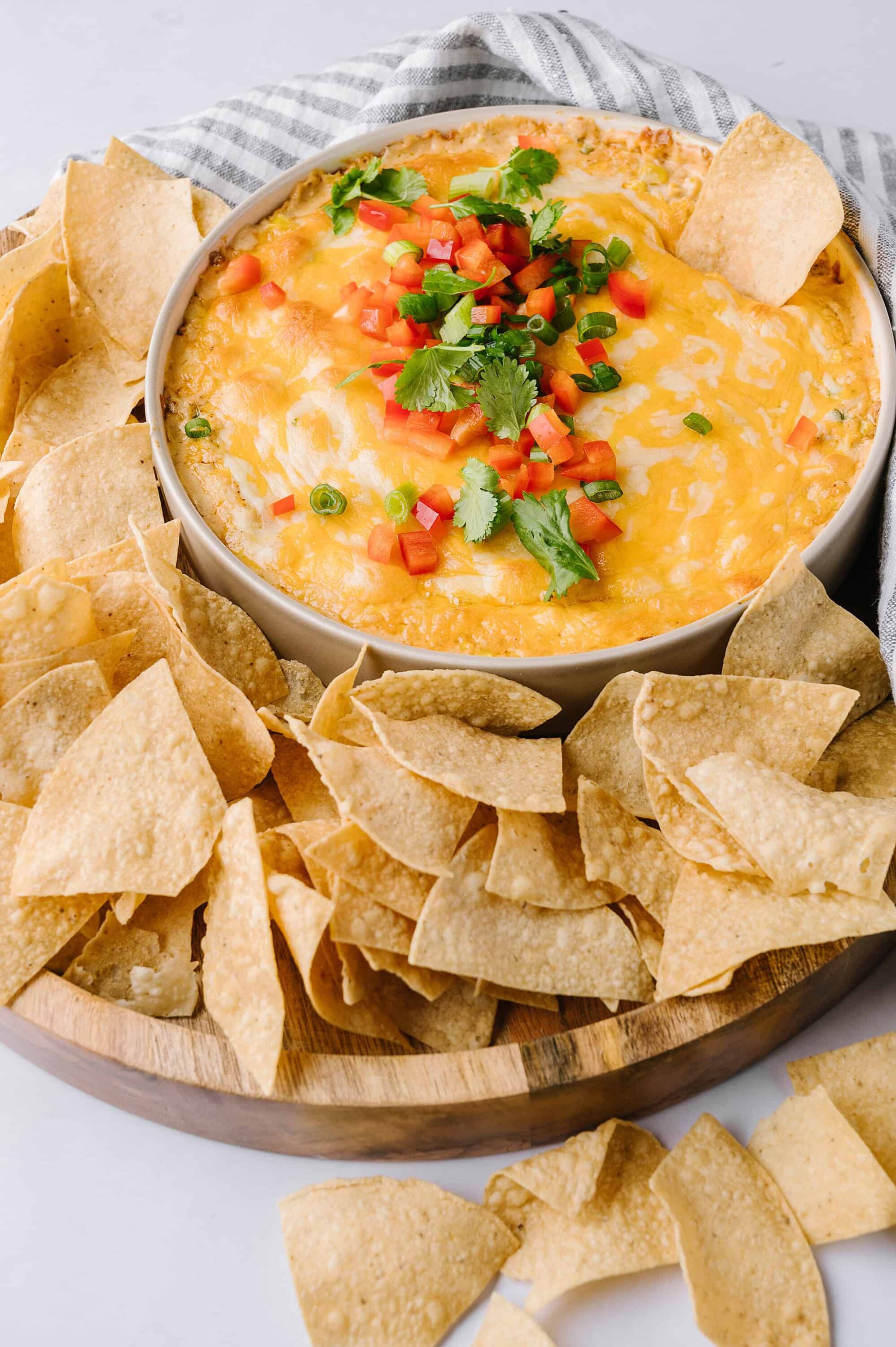 Cheesy nacho dip with diced tomatoes and green onions, served with tortilla chips on wooden platter.