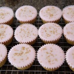 Delicate vanilla cupcakes with pink frosting and gold sprinkles on a cooling rack.