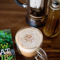 Steamed coffee drink topped with foam and cocoa powder in a glass mug on wooden table.