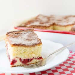 Fluffy cherry crumb cake slice with powdered sugar on white plate and whole cake in background.