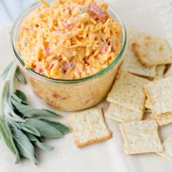 Creamy bacon and cheddar cheese potato salad in a glass bowl with crackers on a white cloth.