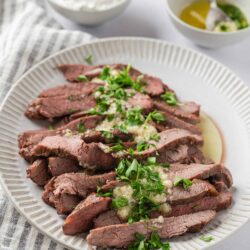 Juicy sliced beef tenderloin with garlic herb butter garnished with fresh parsley on a textured white plate.