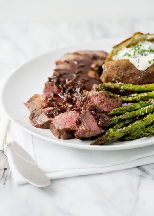 Juicy prime rib roast with gravy, fresh asparagus, baked potato with sour cream on white plate.