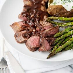 Juicy medium-rare steak with mushroom sauce, baked potato with sour cream, and roasted asparagus on white plate.