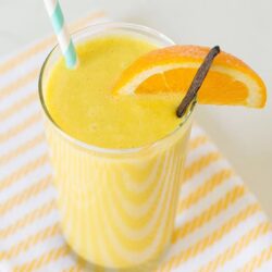 Bright yellow orange juice with fresh orange slices and a vanilla bean, served in a clear glass with a striped straw.