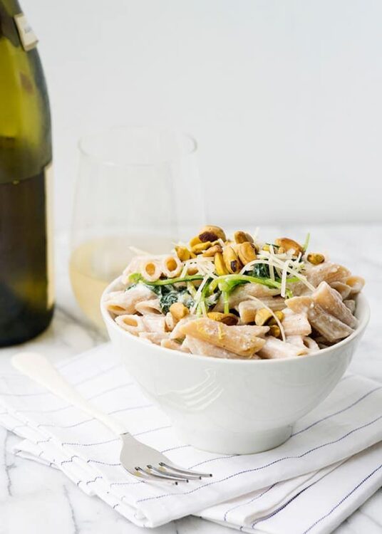 Creamy pasta salad with vegetables and nuts on white bowl, wine glass, and bottle on white table.