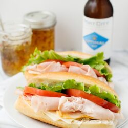 Fresh turkey sandwiches with lettuce, tomato, and cheese on crusty baguette bread, served with condiments and a cold beverage.