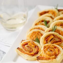 Cream cheese and smoked salmon pinwheels on white platter with garnishes and wine in background.
