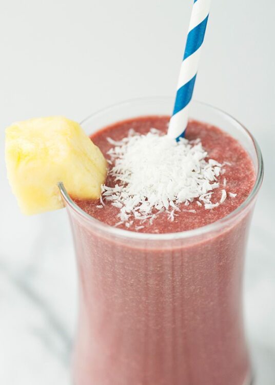 Pineapple strawberry smoothie in a tall glass with shredded coconut and a striped straw.