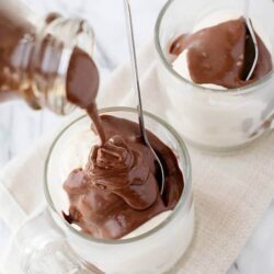 Rich hot fudge being poured over vanilla ice cream in glass mugs.