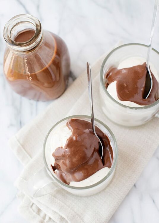 Rich chocolate pudding served in glass cups topped with extra chocolate sauce and a chocolate syrup bottle in the background.
