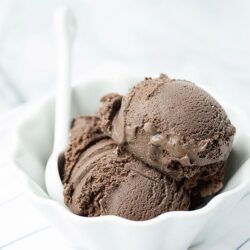 Rich chocolate ice cream scoops in a white bowl with a white spoon on a striped cloth.