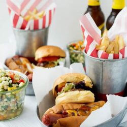 Classic American hot dogs with toppings in a metal tub, picnic style, with fries, beer, and side salad.