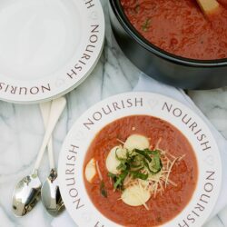 Tender tomato basil soup garnished with fresh basil, mozzarella, and grated cheese on a "Nourish" plate.