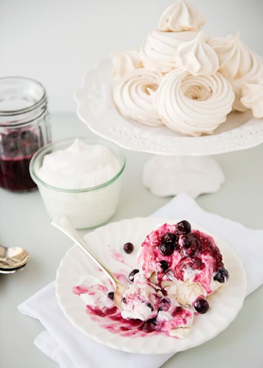 Fluffy meringue cookies with whipped cream and berry sauce dessert on a white cake stand.